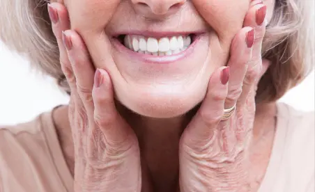 Eine ältere Frau mit strahlendem Lächeln hält ihre Wange, symbolisch für Freude und Zufriedenheit mit ihrer Zahngesundheit.