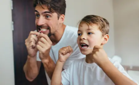 Ein Mann und ein Junge, vermutlich Vater und Sohn, lächeln und putzen sich gemeinsam die Zähne vor einem Badezimmerspiegel.